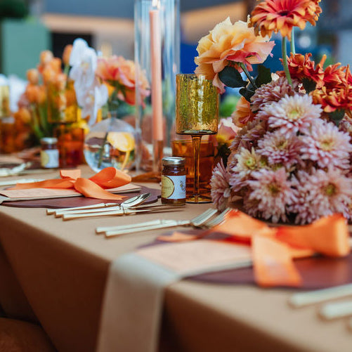A long banquet table set in a large event hall, featuring floral centerpieces with orange, pink, and yellow flowers, gold goblets, and place settings under a clear marquee roof.