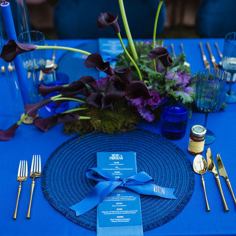A styled event table setting with a bright blue tablecloth, matching blue place settings, woven round placemats, blue glassware, gold cutlery, and a floral centerpiece with dark calla lilies.