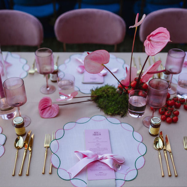 A styled pink and blush table setting with scalloped white placemats, gold flatware, pink napkins and candlesticks, pink flowers, and greenery on a beige tablecloth.